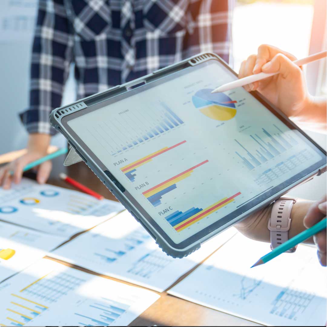 In a sunlit office, one person holds a pen and points at a tablet displaying financial data, while another person—standing on the opposite side of the table, wearing a blue plaid flannel shirt—also holds a pen and reviews financial documents.