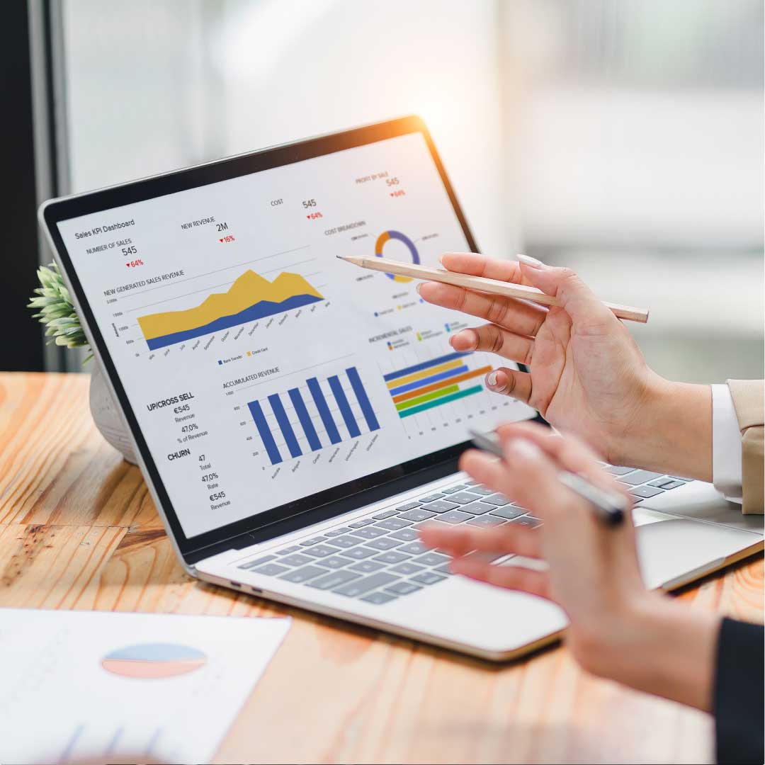 A close-up image of two coworkers' hands holding pens and pointing at financial data displayed on a laptop.