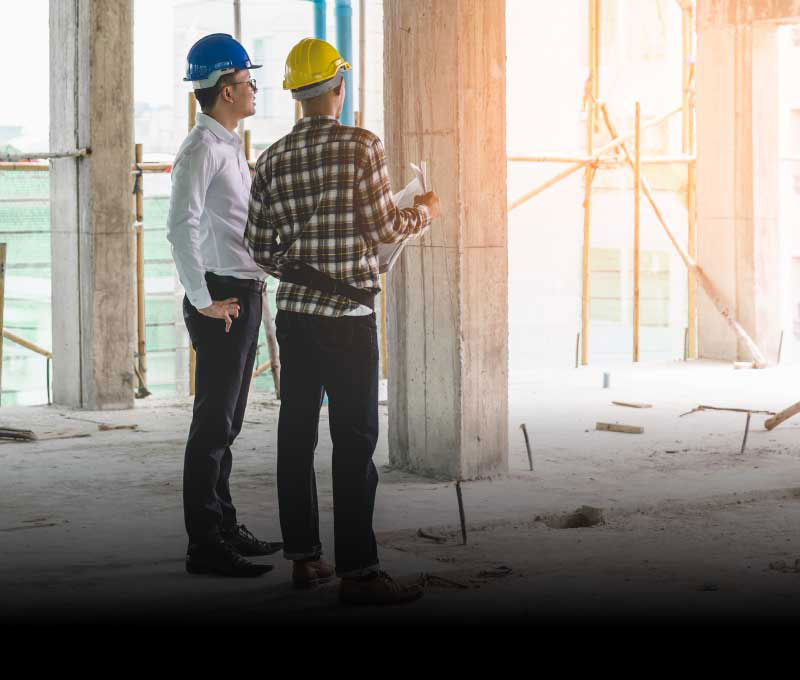 Two men wearing hard hats are looking off into the distance while standing in a construction site, one is holding architectural plans.